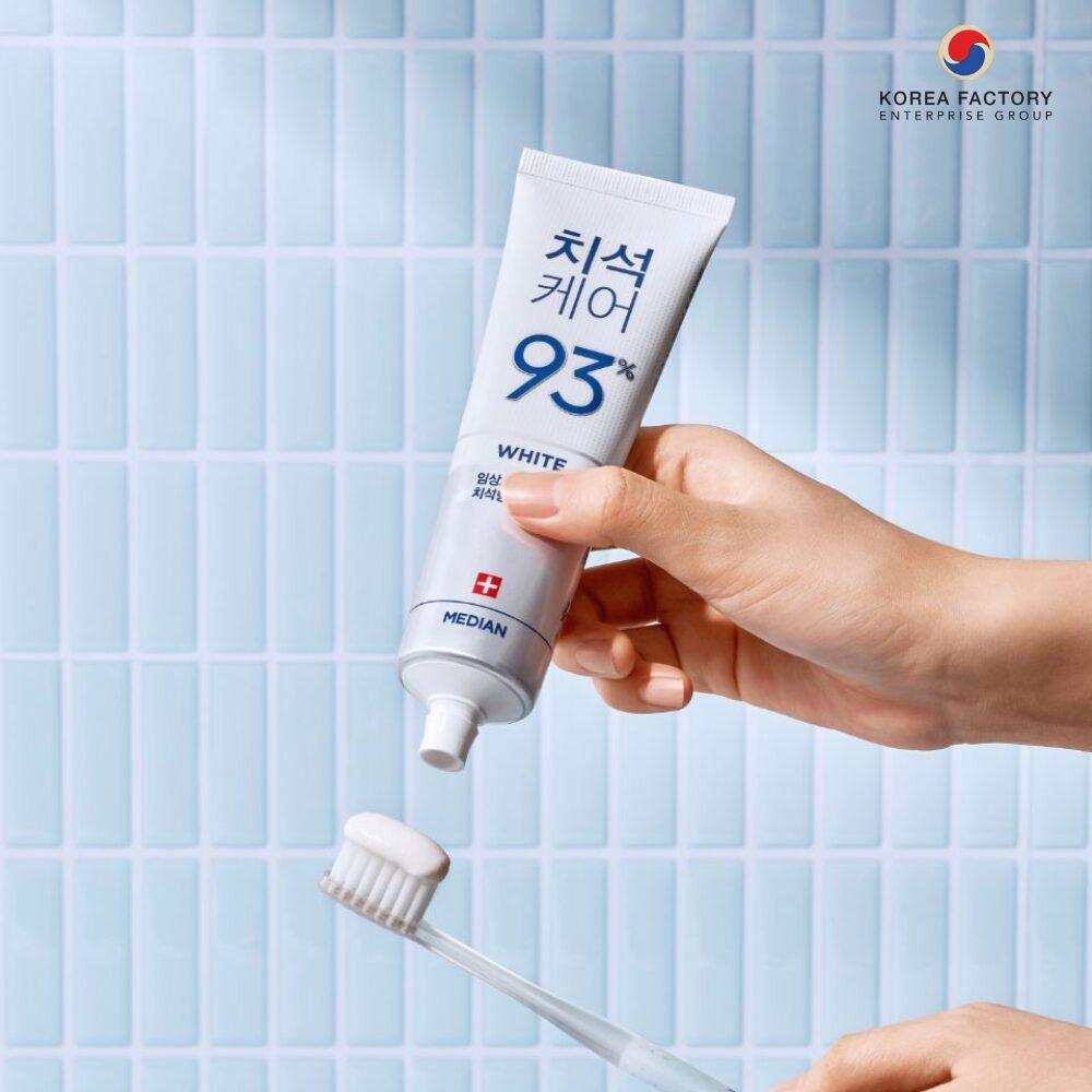 มือของผู้ใช้กำลังถือหลอดผลิตภัณฑ์ดูแลช่องปากสีขาว บีบเนื้อผลิตภัณฑ์ลงบนแปรงสีฟันสีขาว ฉากหลังเป็นกระเบื้องห้องน้ำสีฟ้าพาสเทล พร้อมโลโก้ Korea Factory ด้านบนซ้ายของภาพ