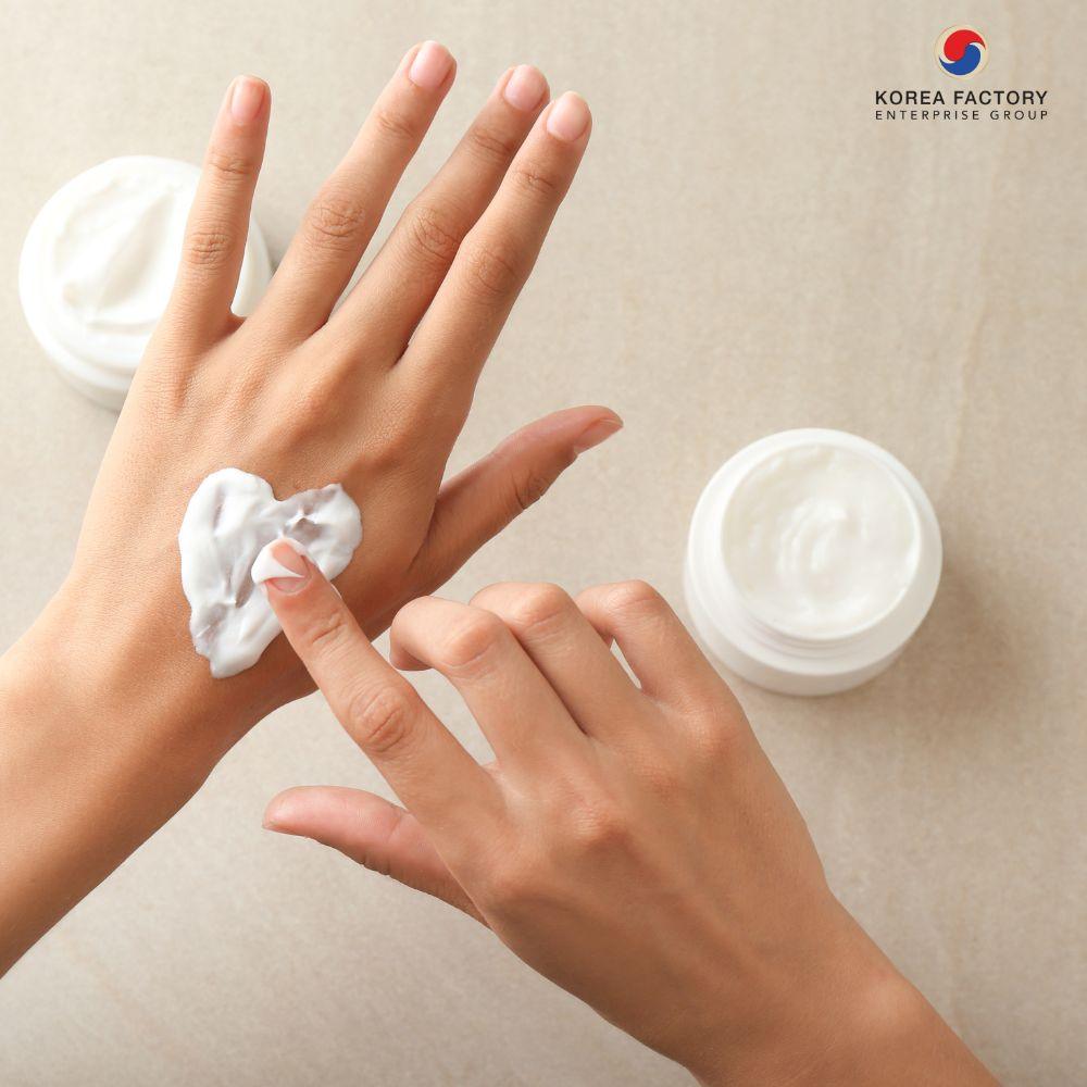 มือผู้หญิงกำลังทาแฮนด์ครีมสีขาวบนหลังมือ มีกระปุกครีมสีขาววางอยู่ด้านข้าง พร้อมกรอบข้อความเกี่ยวกับ HAND CREAM และบริการโรงงานรับผลิตแฮนด์ครีม CosKorea Group