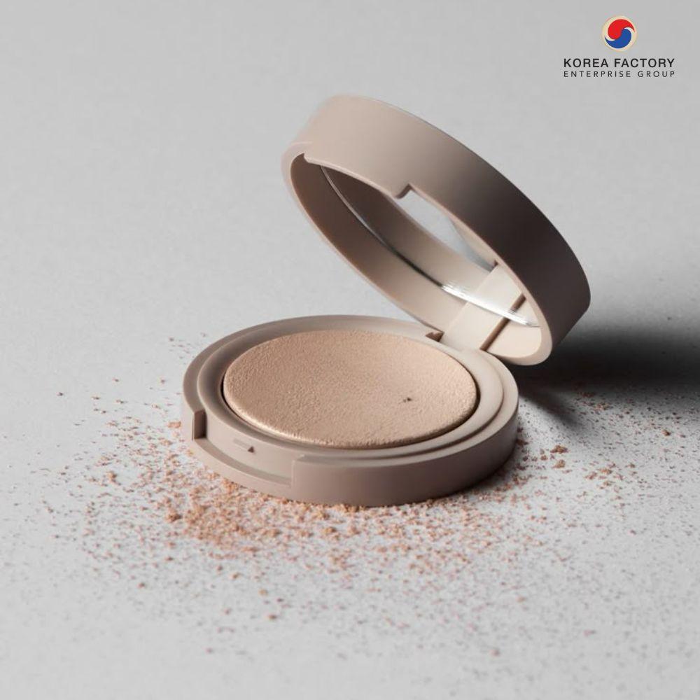 ตลับแป้งอัดแข็งสีนู้ดฝาเปิด วางบนพื้นสีเทาพร้อมผงแป้งกระจาย และโลโก้ Korea Factory Enterprise Group